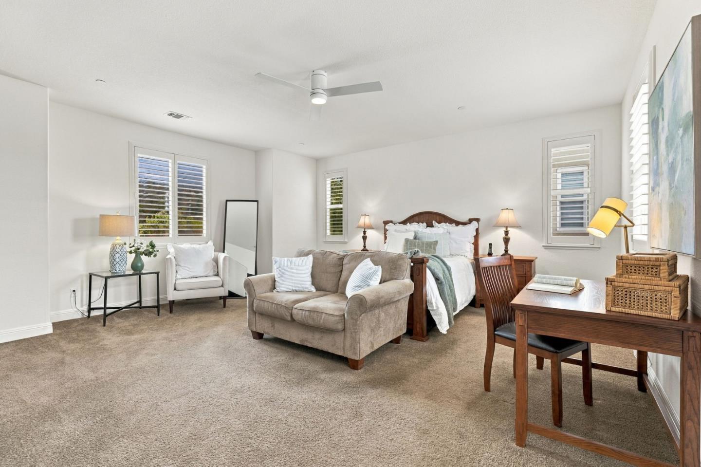 Staged bedroom with a wood-framed bed, loveseat, and writing desk beneath shuttered windows.