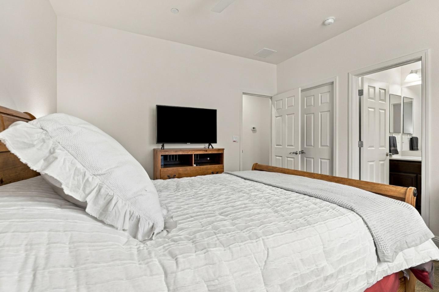 Furnished bedroom with white bedding, a wooden bed, and a television beside the bathroom doorway.