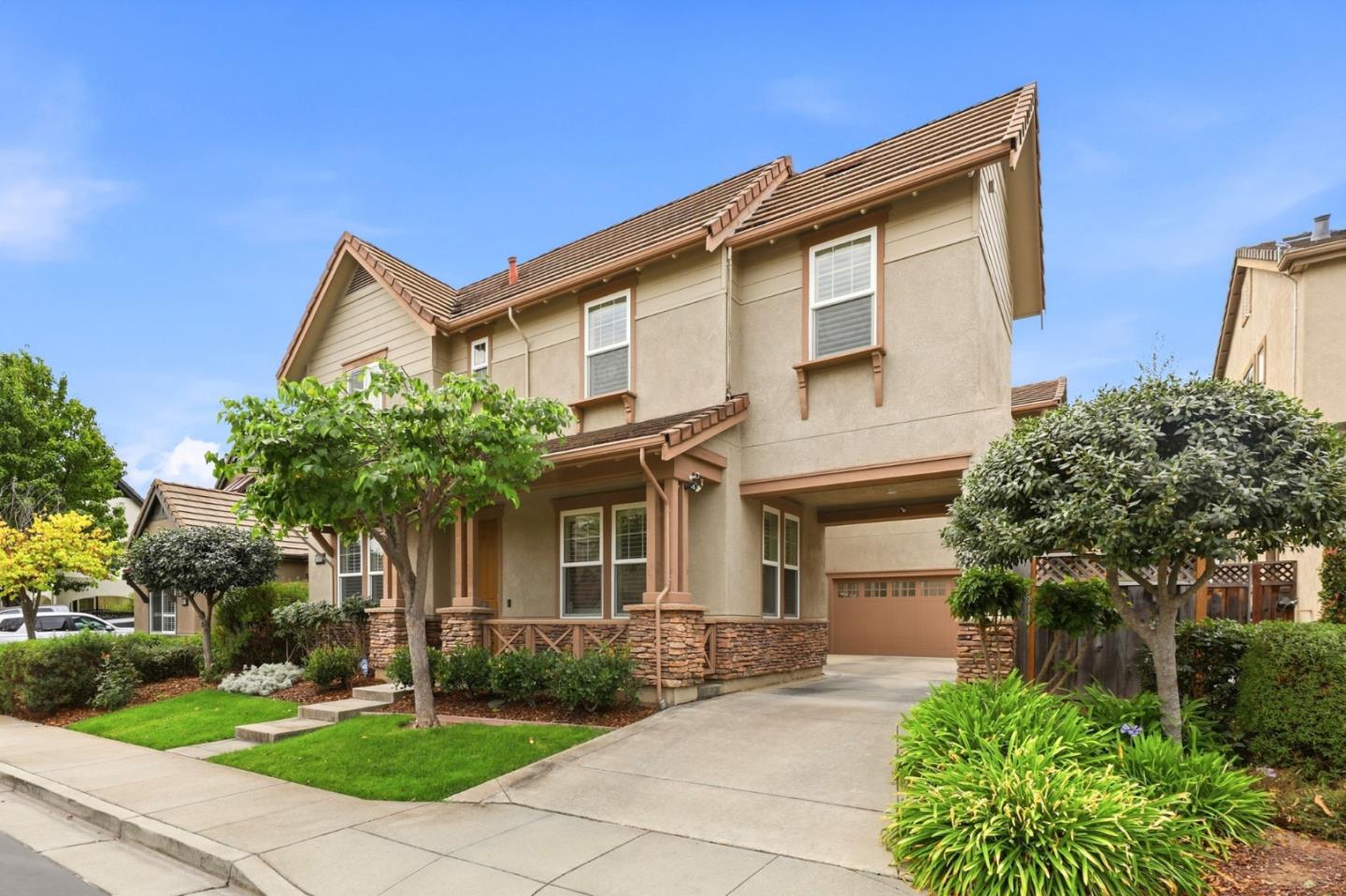 Two-story beige house with brown tile roofs, stone accents, landscaped lawn, and attached garage.