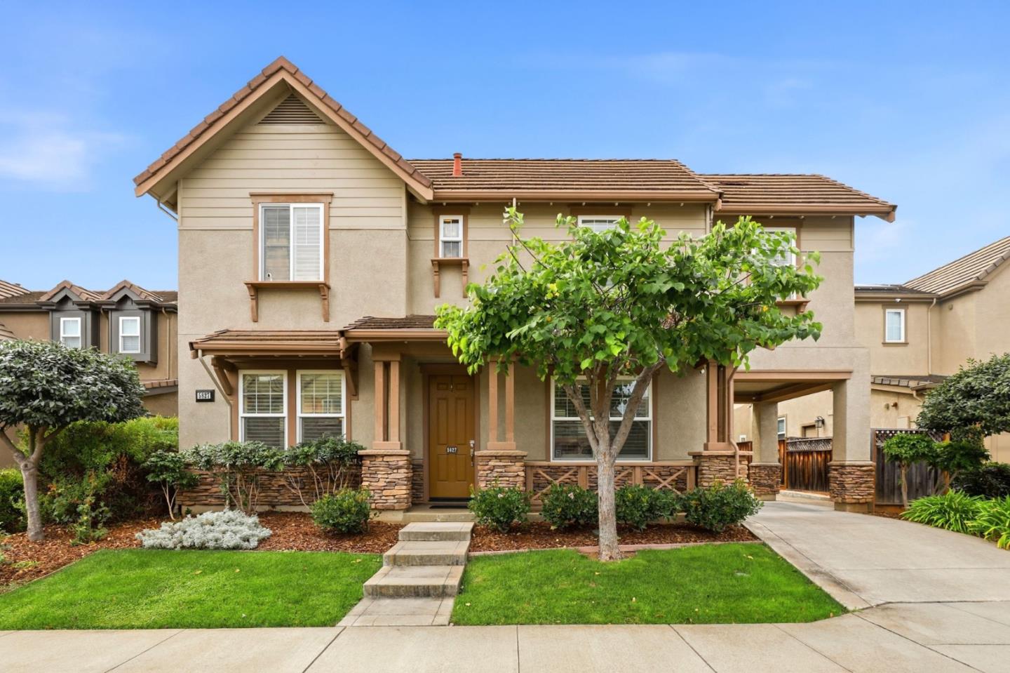 Two-story beige home with a tiled roof, landscaped front lawn, and a leafy tree beside the entrance.