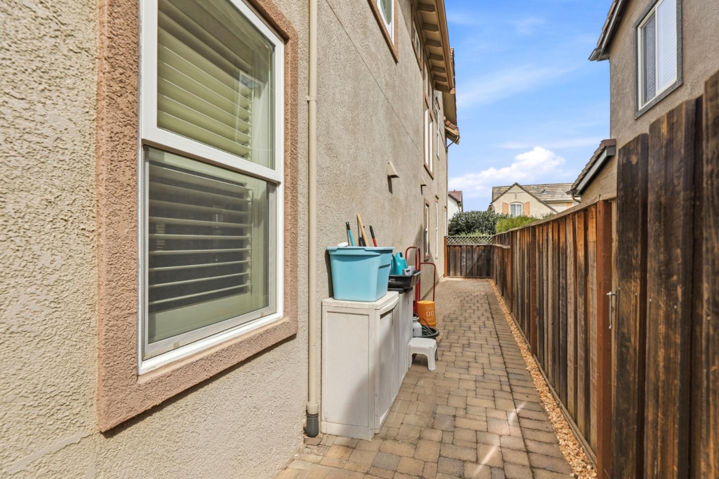 Paved side yard beside a stucco house, with a tall wooden fence and storage bins.