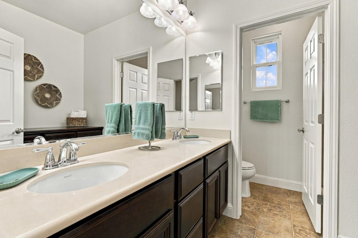 Bathroom with a double-sink vanity, dark wood cabinets, turquoise towels, and a window beside the toilet.