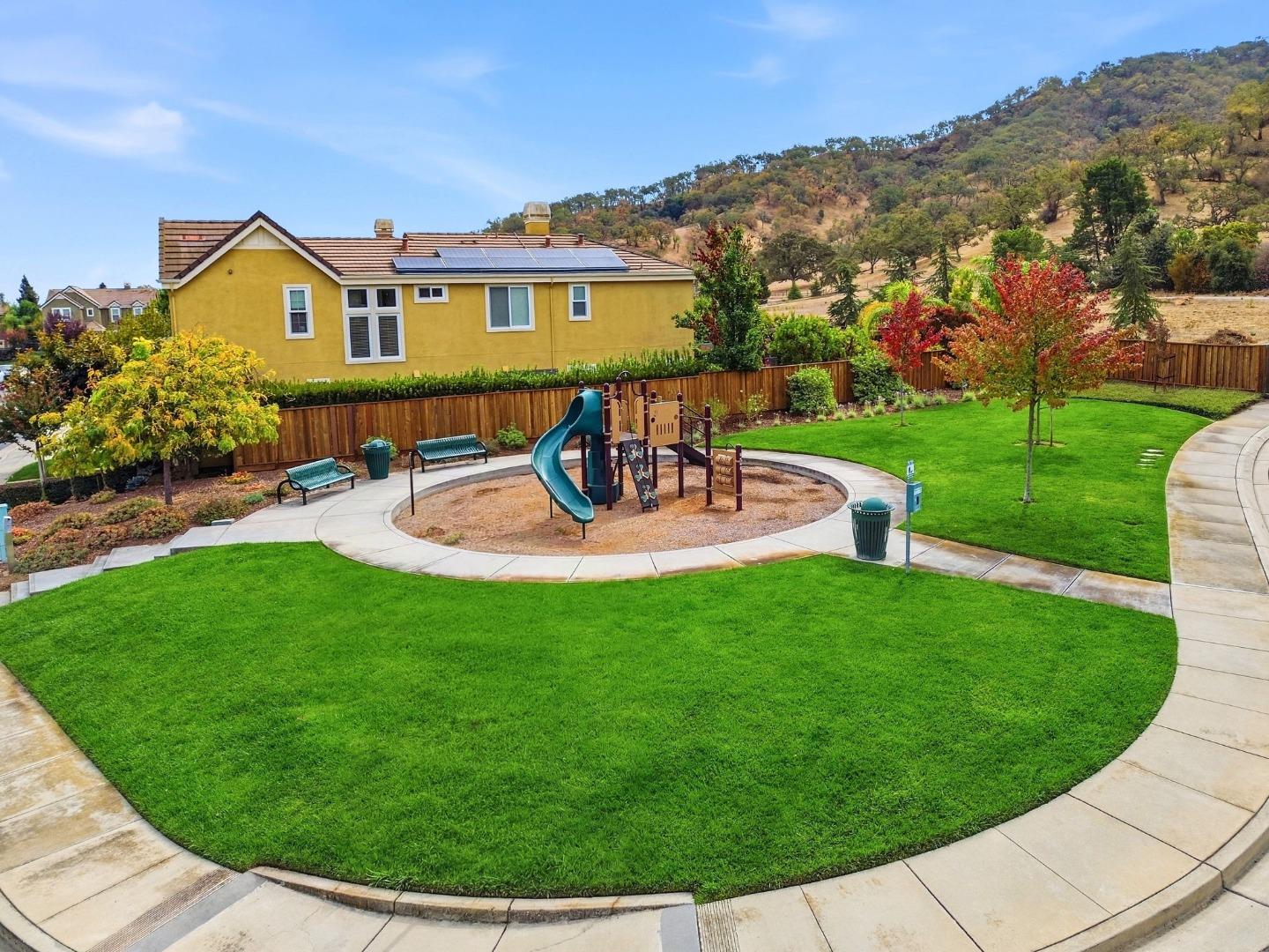 Community playground with teal slide, circular path, green lawn, and hillside behind a wooden fence.