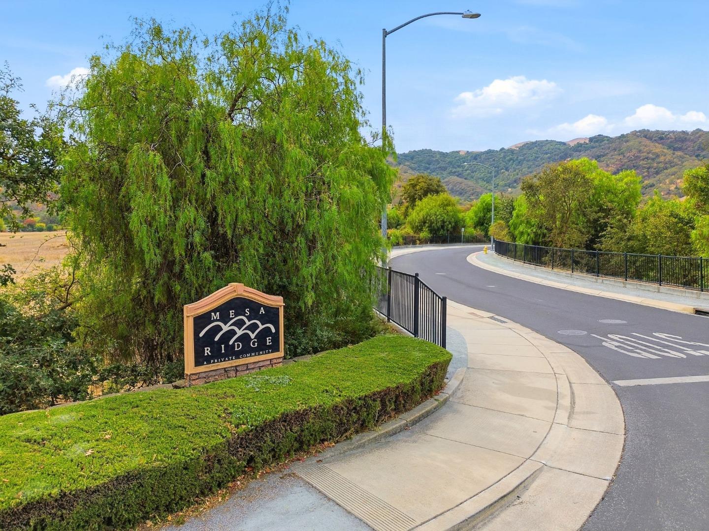 Mesa Ridge community entrance sign beside a curving road, with green hills beyond.
