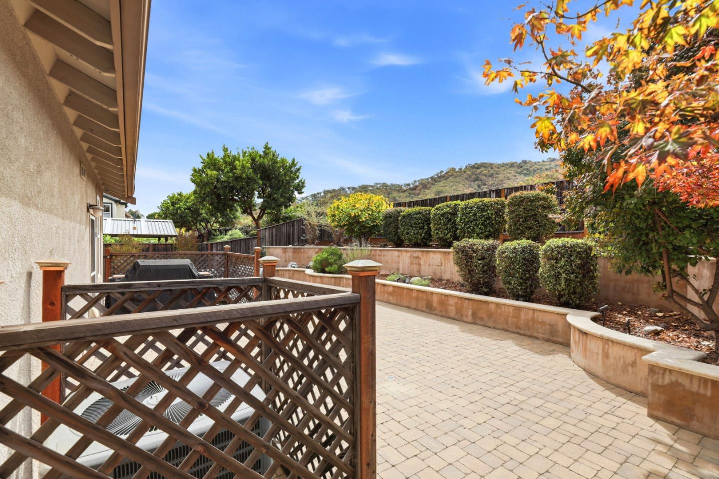 Paved patio with a lattice railing, raised planters, and a wooded hillside beyond the yard.