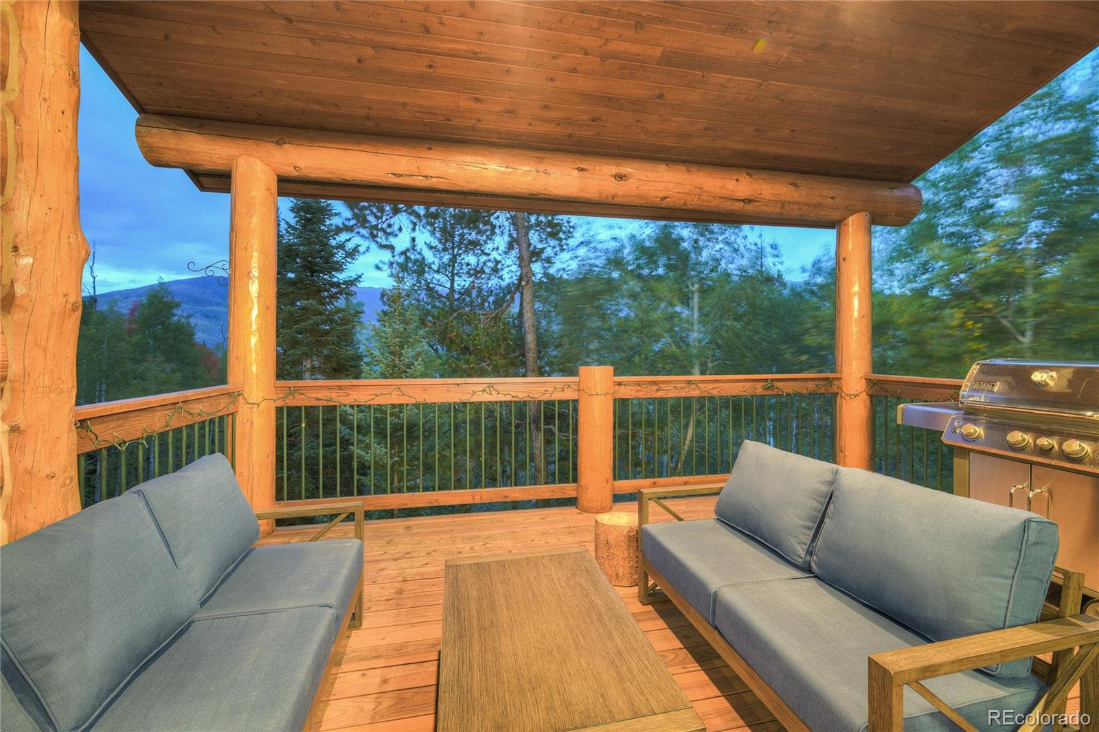 Covered log deck with blue seating and grill, overlooking trees and a distant mountain.