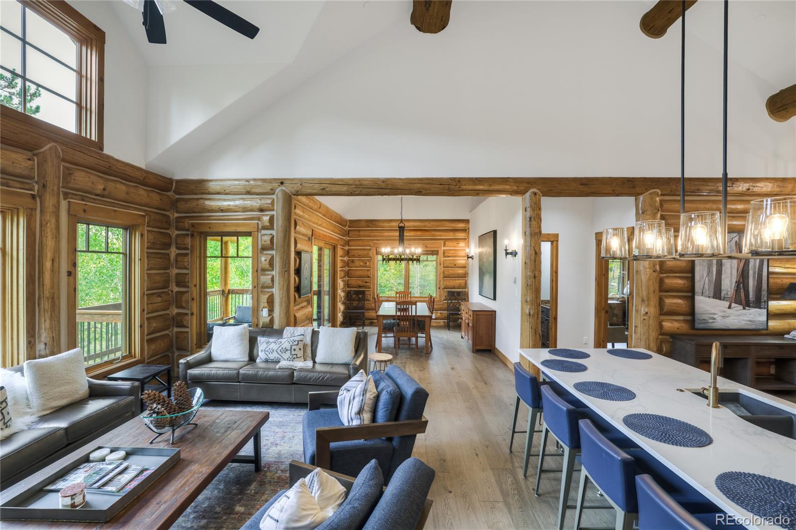 Open-plan log great room with vaulted ceiling, timber beams, and a kitchen island lined with blue stools.