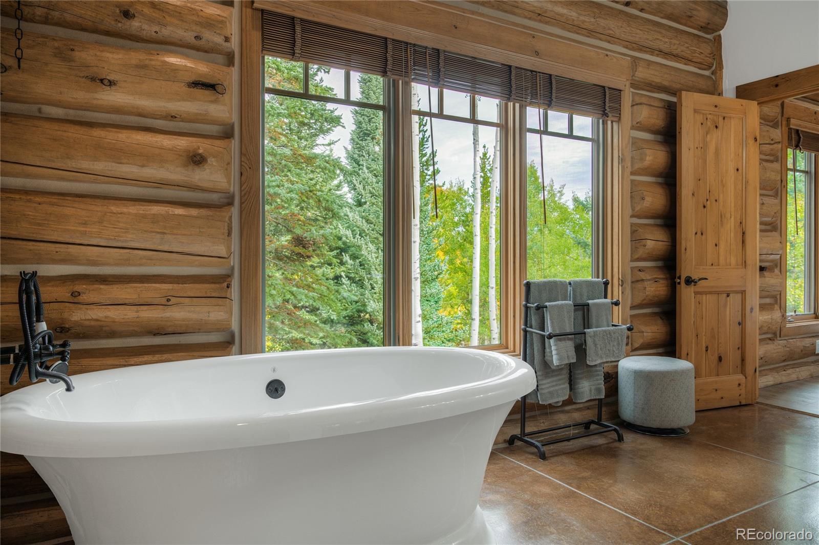 Freestanding bathtub in a log-cabin bathroom with large windows overlooking trees.