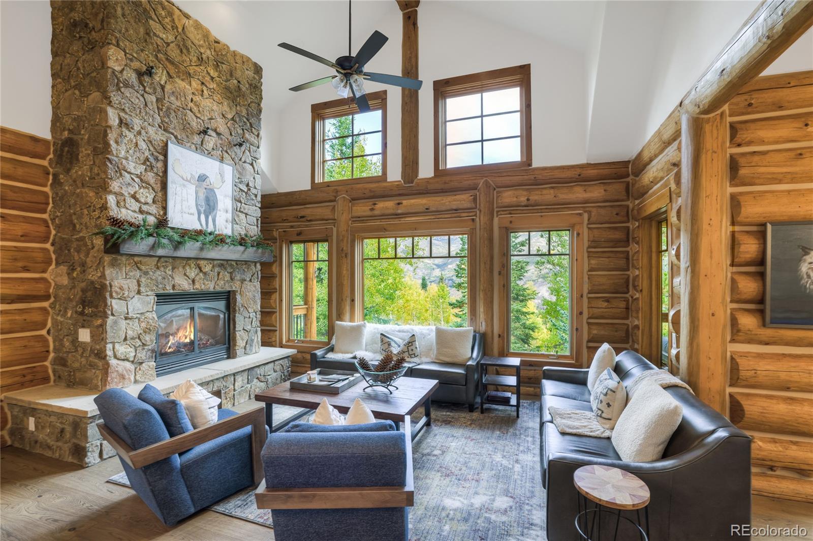 Log home living room with a stone fireplace, timber beams, and mountain views through large windows.