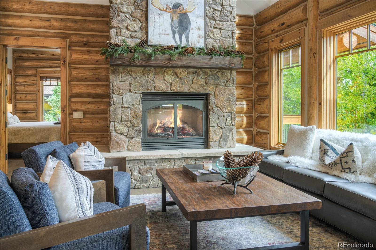 Log-cabin living room with a stone fireplace, exposed timber walls, and tall windows.