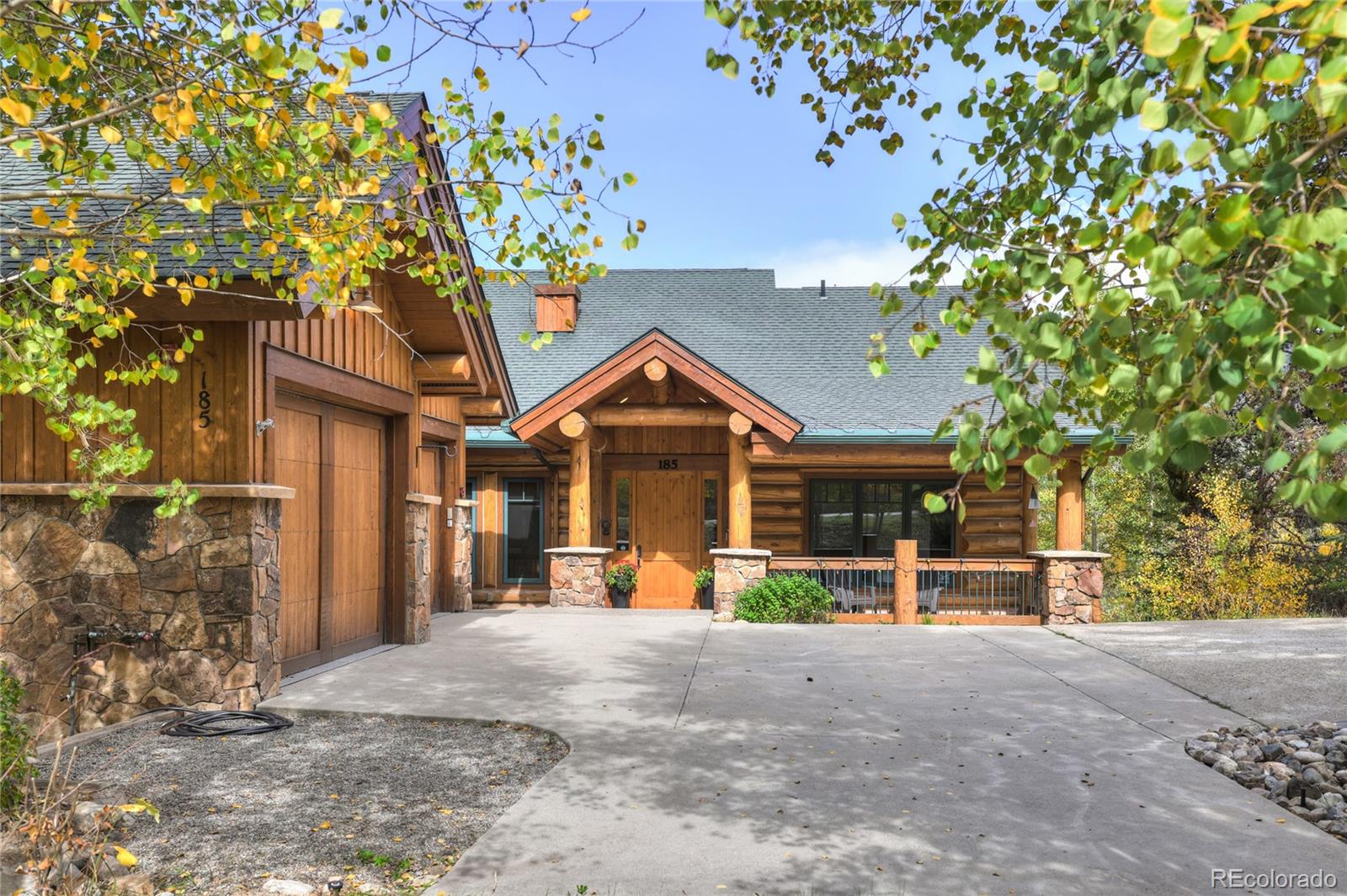 Rustic wood-and-stone home with a covered entrance and broad driveway, framed by autumn leaves.