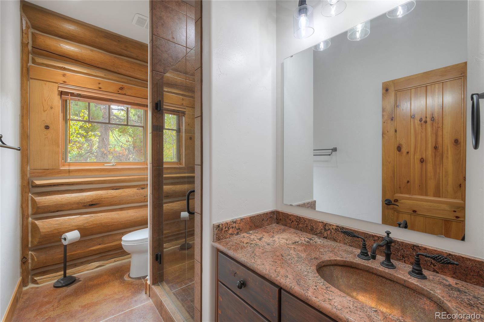 Rustic log-cabin bathroom with glass shower, wood-framed window, and granite-topped vanity.