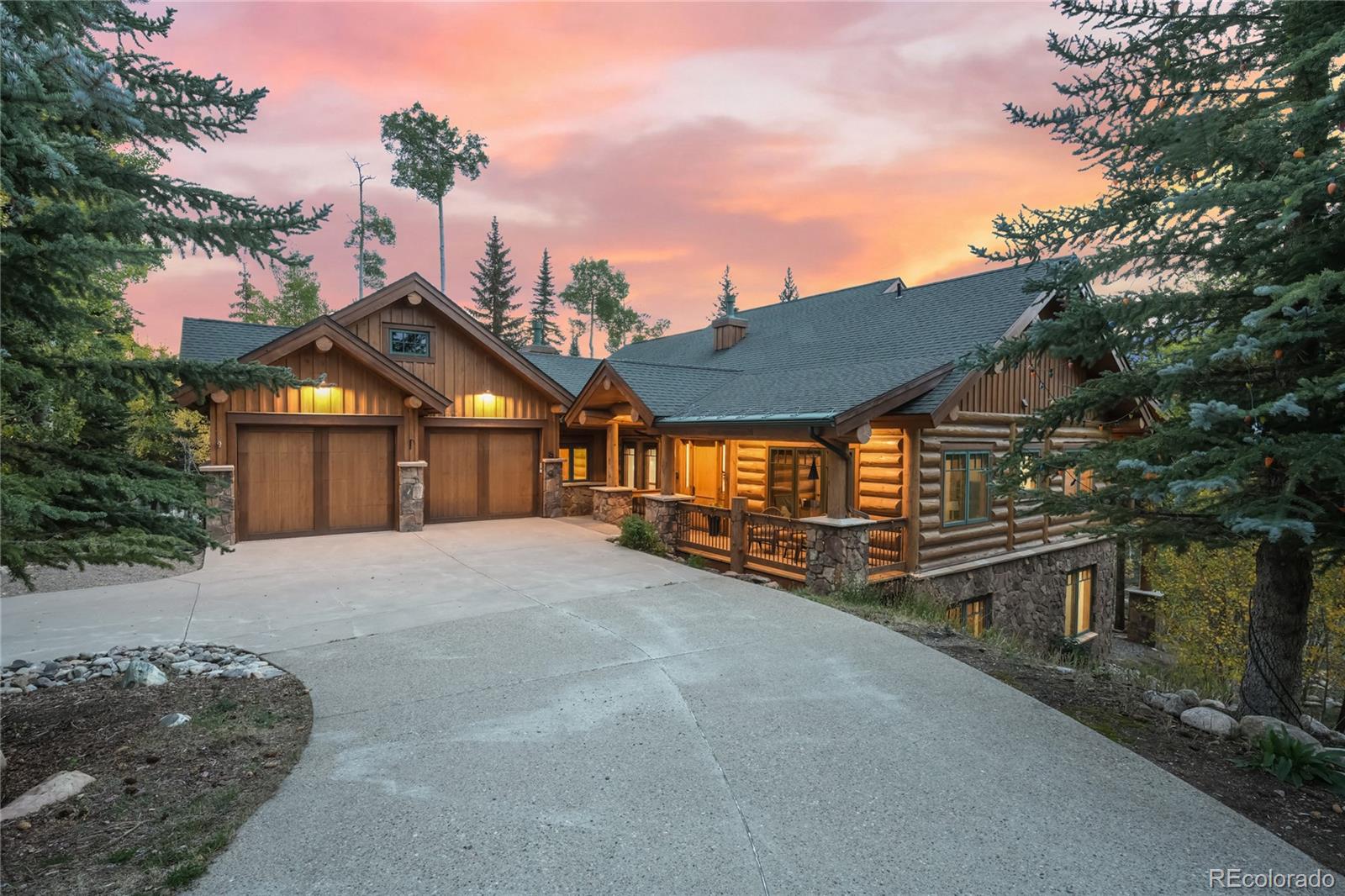 Rustic log home with a two-car garage and stone foundation, framed by evergreens at dusk.