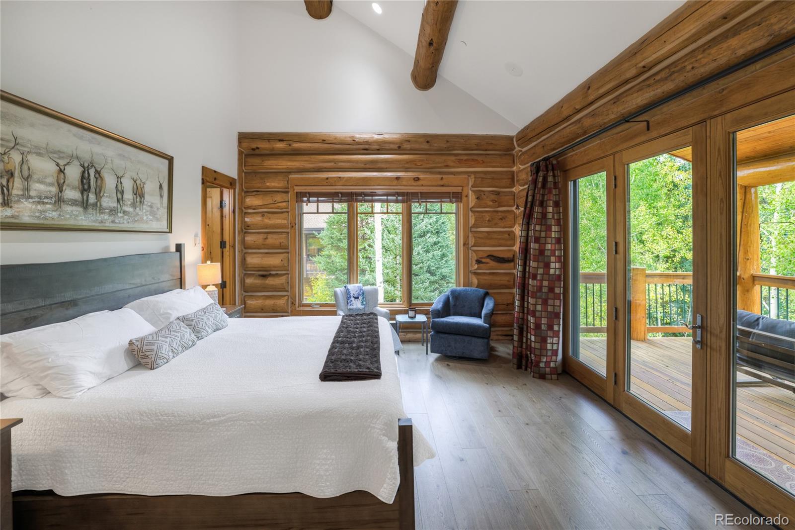 Bedroom with rustic log walls, exposed beams, and glass doors opening to a tree-lined deck.