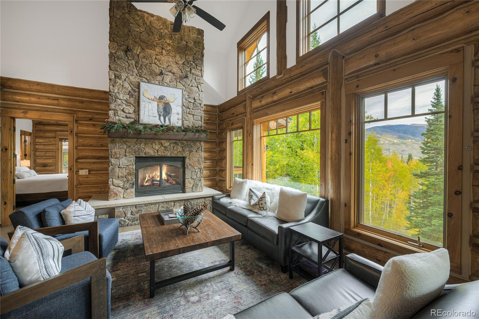Log-cabin living room with a stone fireplace and large windows overlooking autumn trees and mountains.