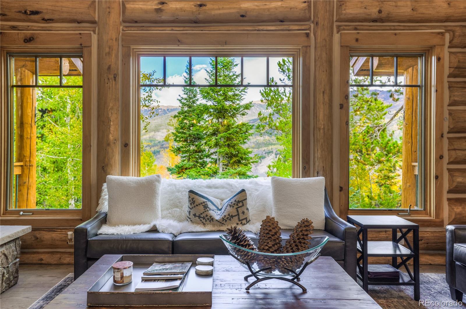 Log-cabin living room with a leather sofa and pinecone centerpiece, facing windows overlooking trees and mountains.