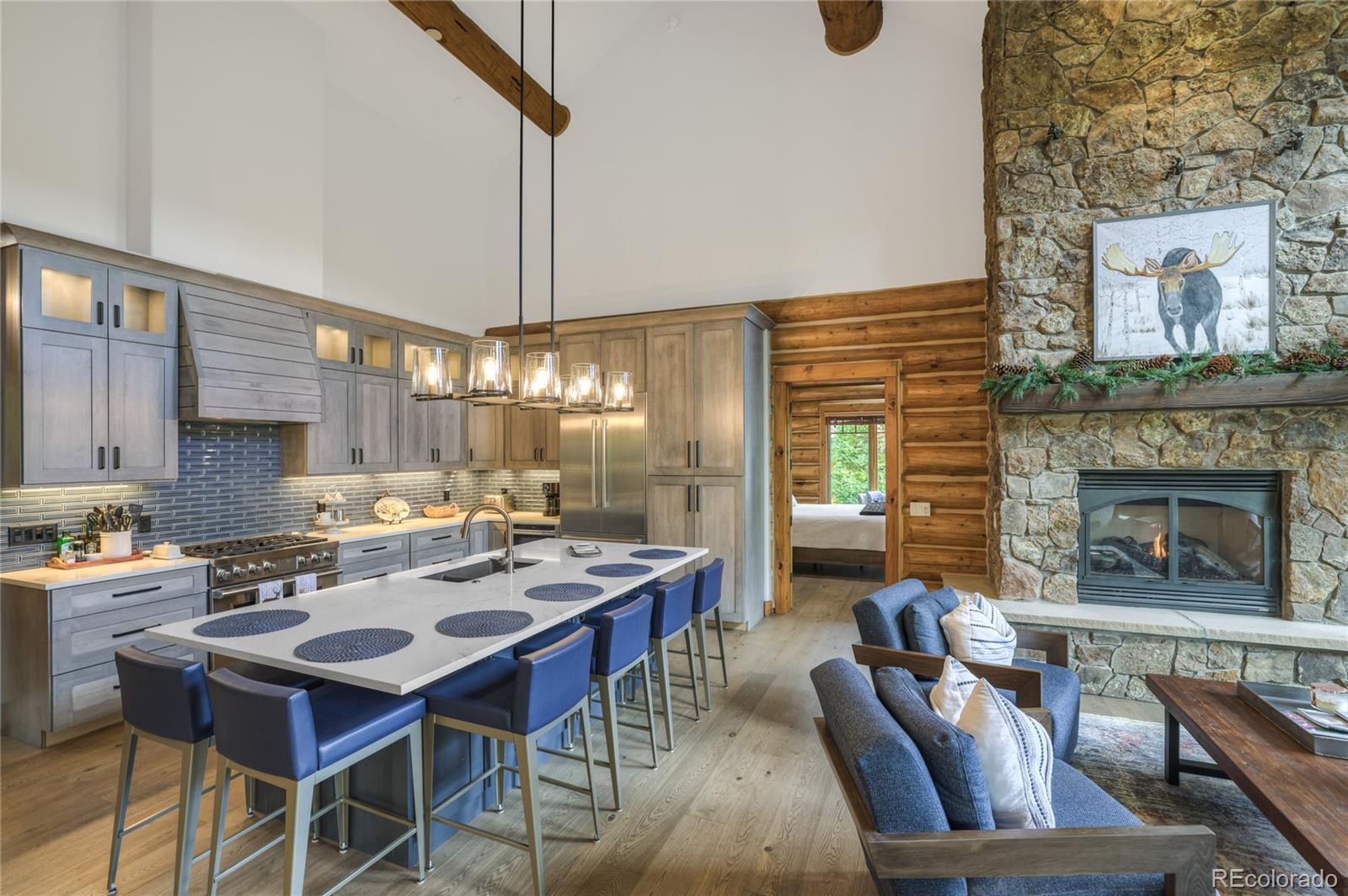 Open kitchen and living area with a white island, blue stools, log walls, and a tall stone fireplace.