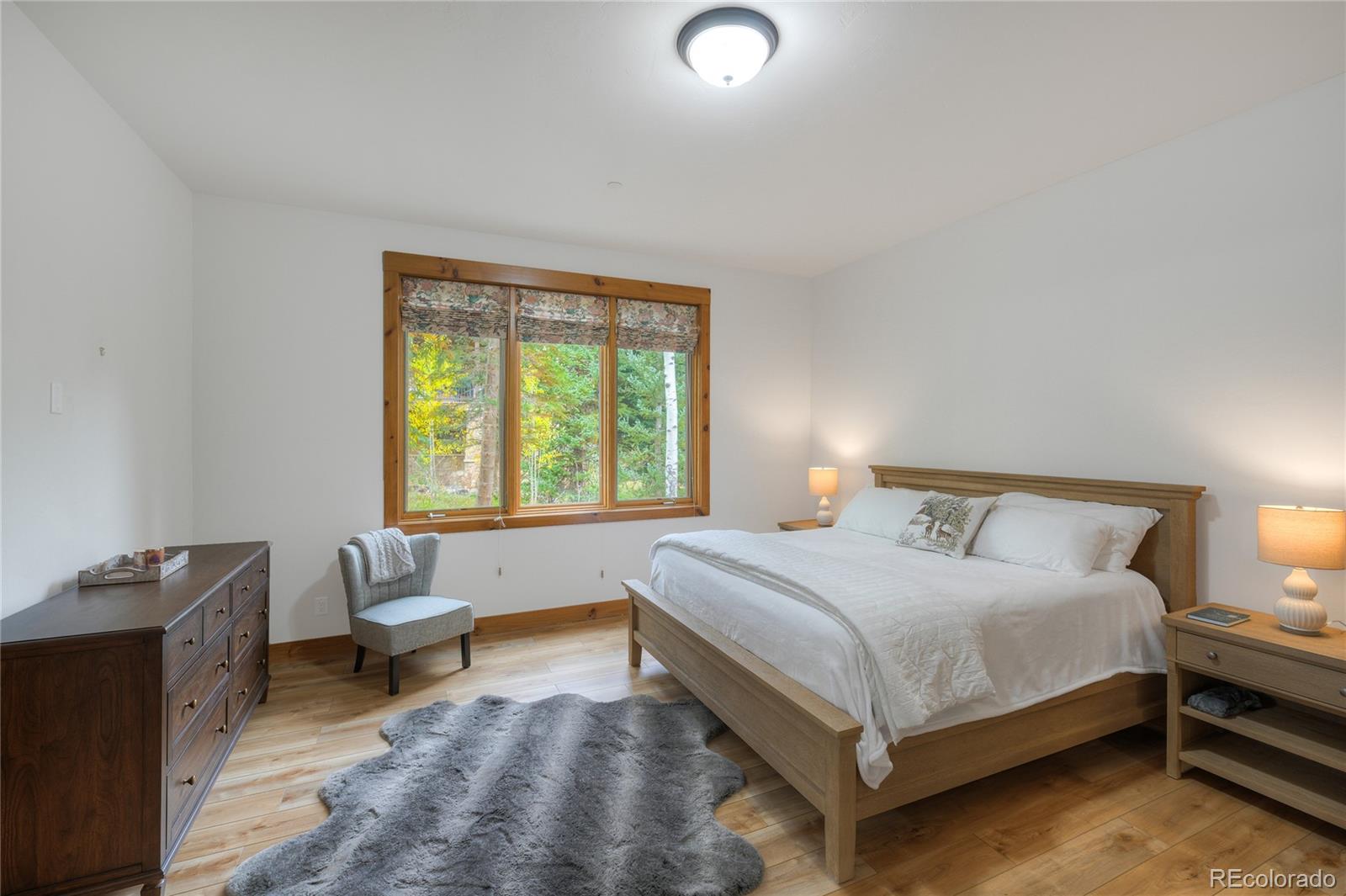 Bedroom with a wooden bed, white linens, wood flooring, and a large window overlooking trees.