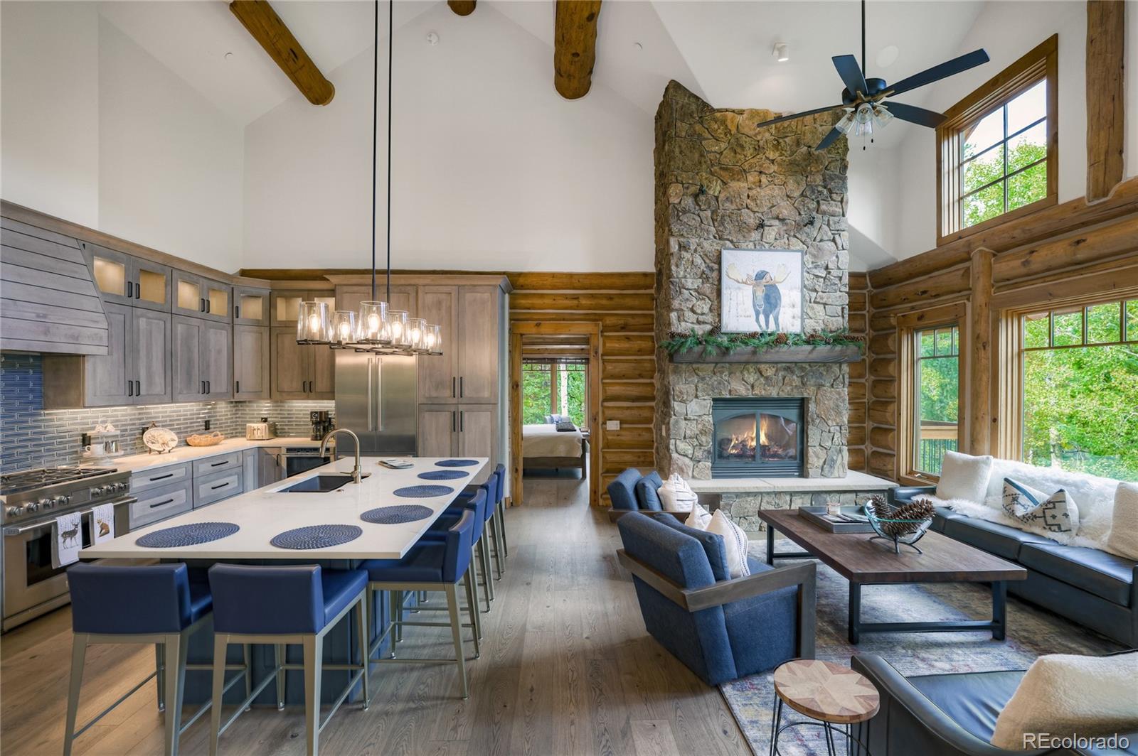 Open-plan kitchen and living room with a stone fireplace, blue island stools, and tall windows.