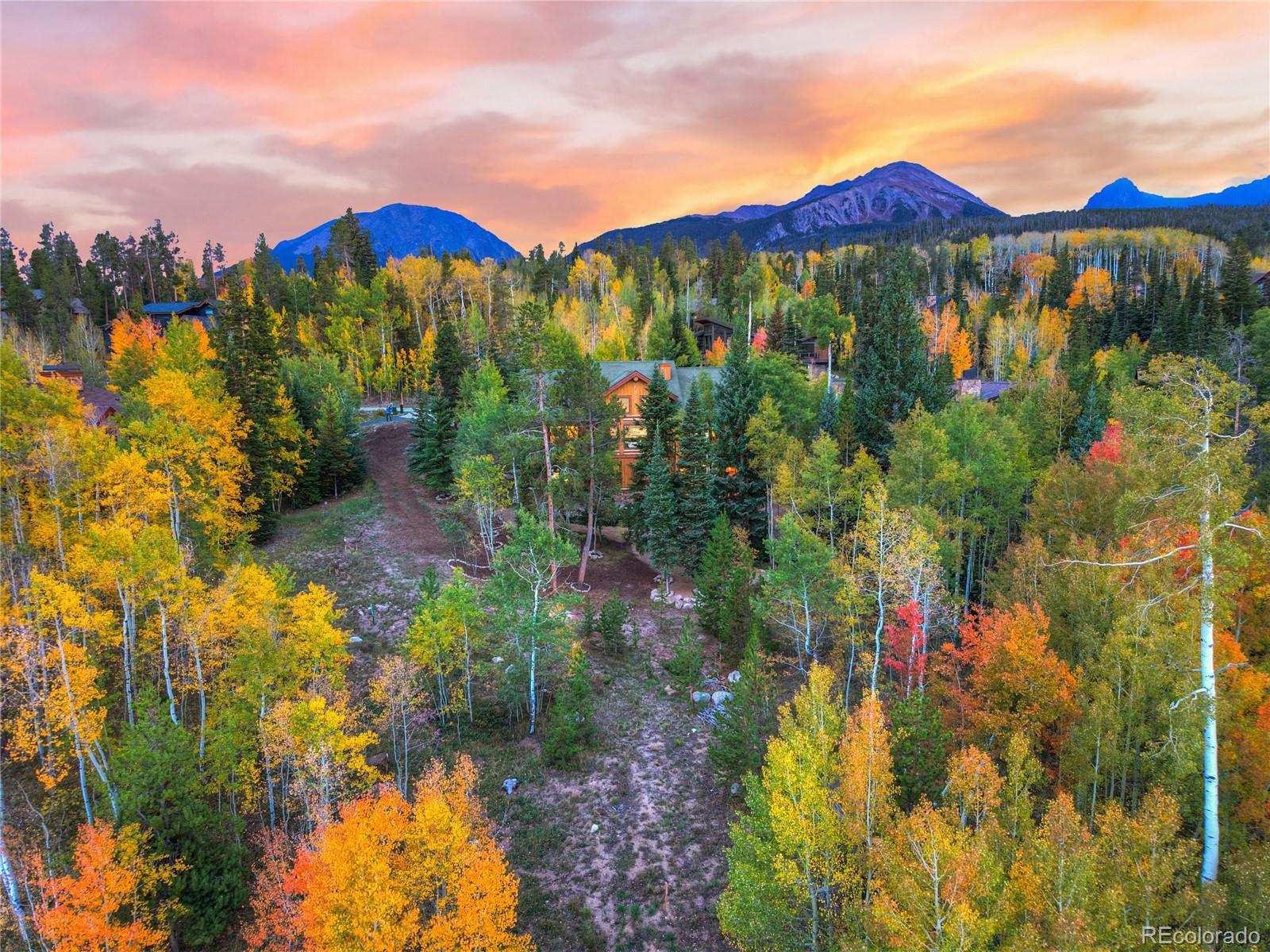 A mountain home nestled among autumn aspens and evergreens beneath a pink-orange sunset sky.