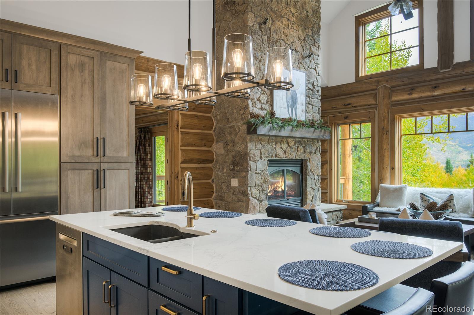 Open-plan kitchen and living room with a white island, stone fireplace, timber walls, and mountain views.