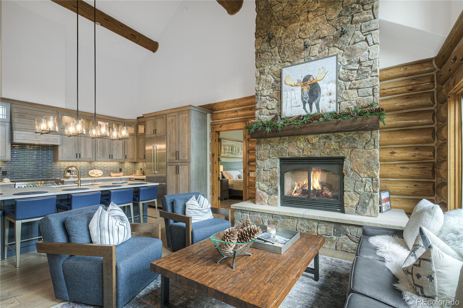 Open-plan living room and kitchen with a stone fireplace, log walls, and blue seating.