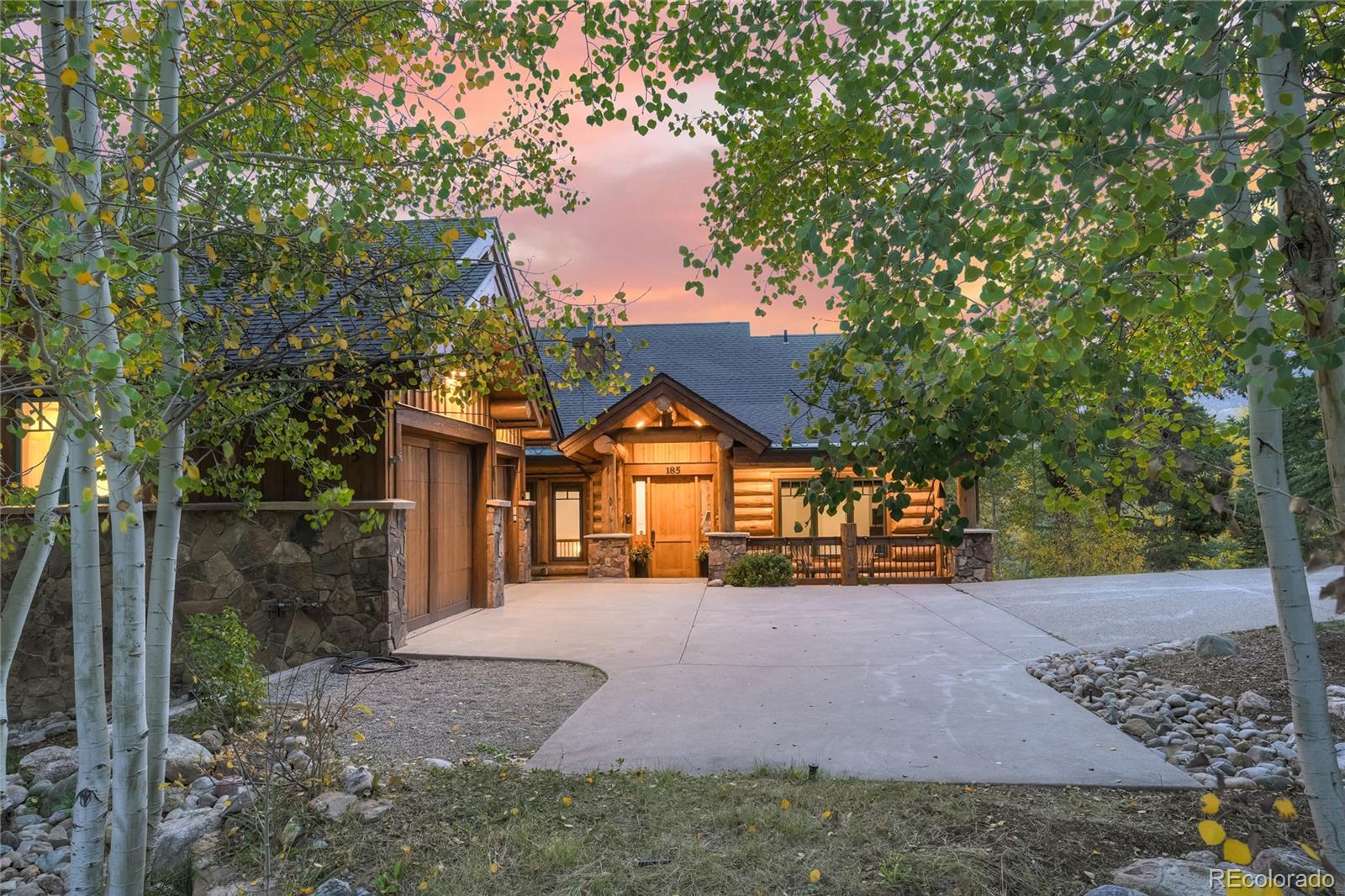 Log-and-stone home with a broad driveway framed by autumn aspens at dusk.