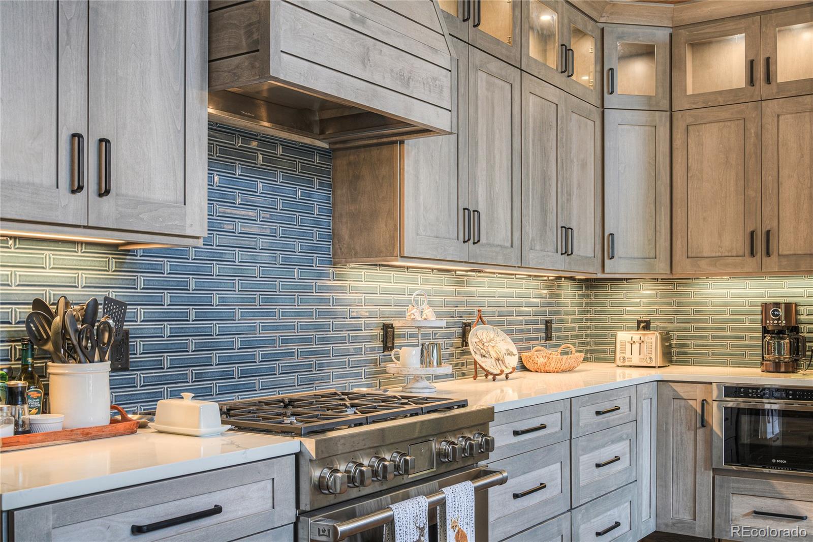 Kitchen with gray wood cabinets, blue-green tile backsplash, white counters, and a large stainless steel range.