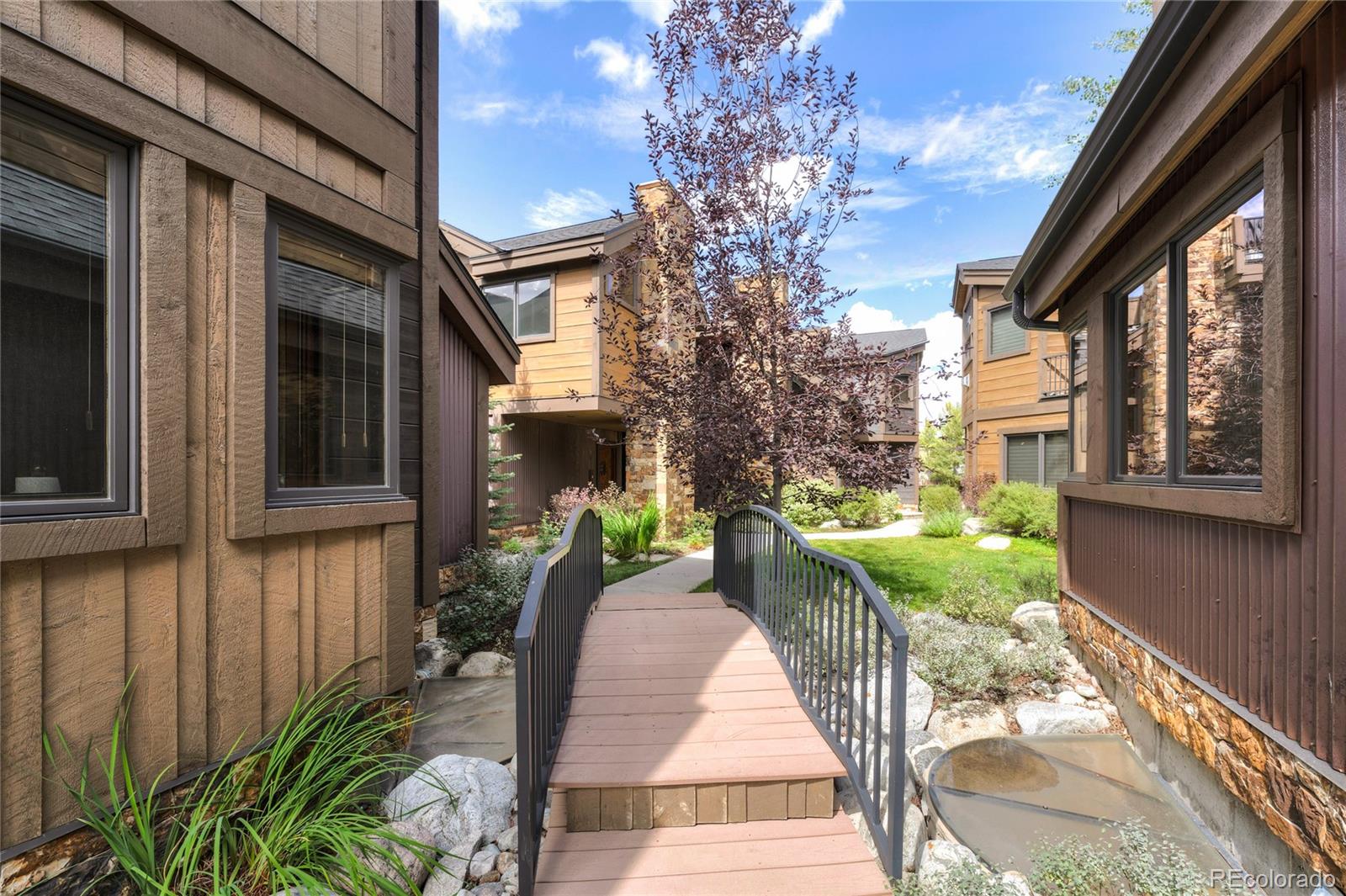 Residential courtyard with a footbridge over a rock-lined water feature between wood-sided buildings.