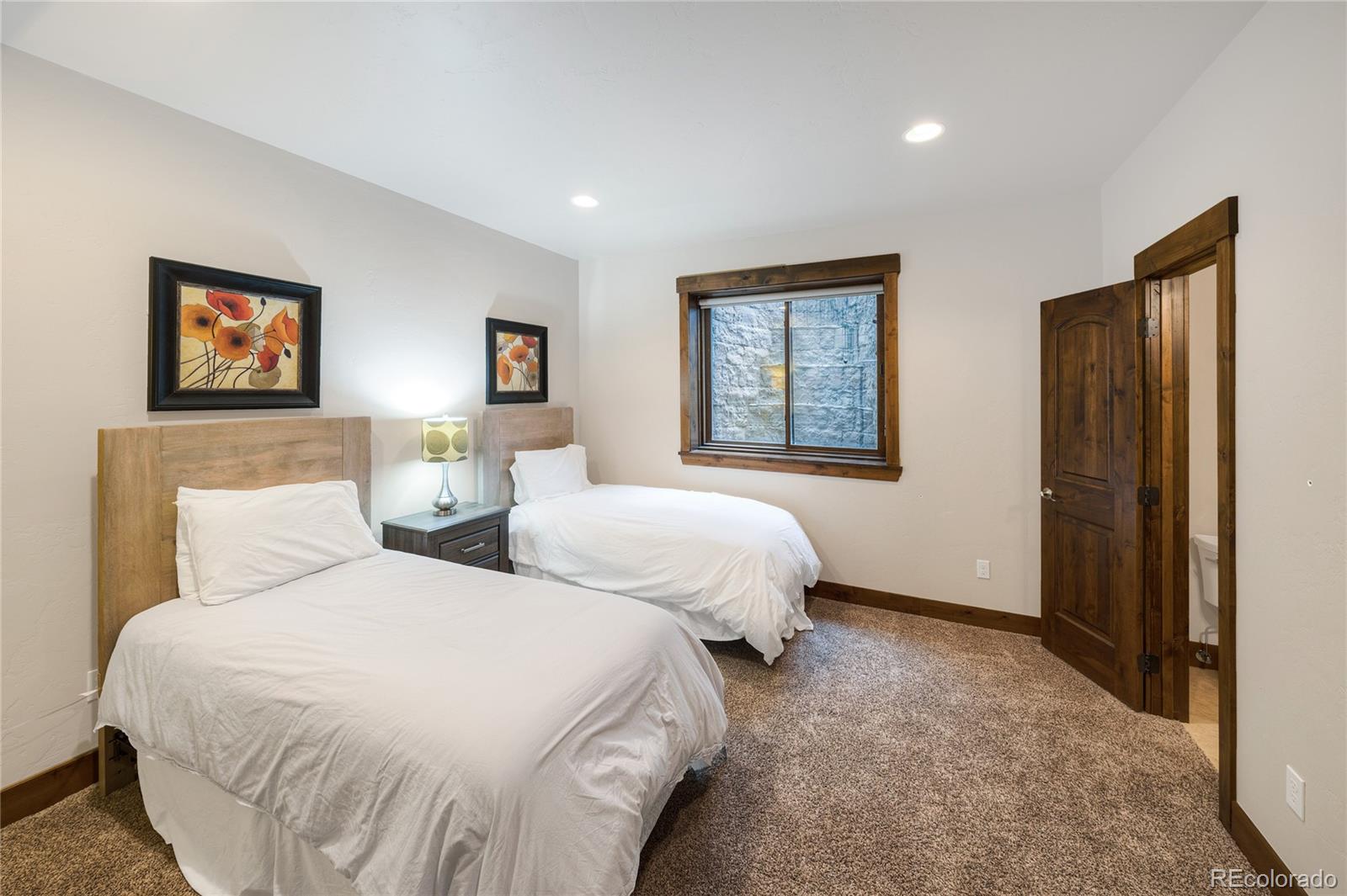 Bedroom with two white-covered beds, wood trim, and a window overlooking a stone wall.