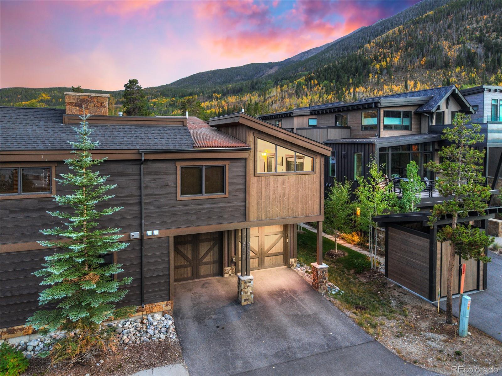 Modern timber-sided mountain home with a garage, framed by autumn foliage and a pink sunset sky.
