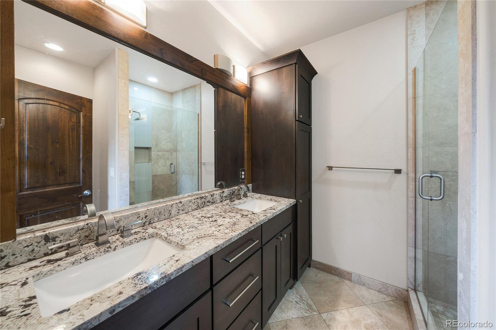 Bathroom with a granite double vanity, dark wood cabinetry, and a glass-enclosed shower.