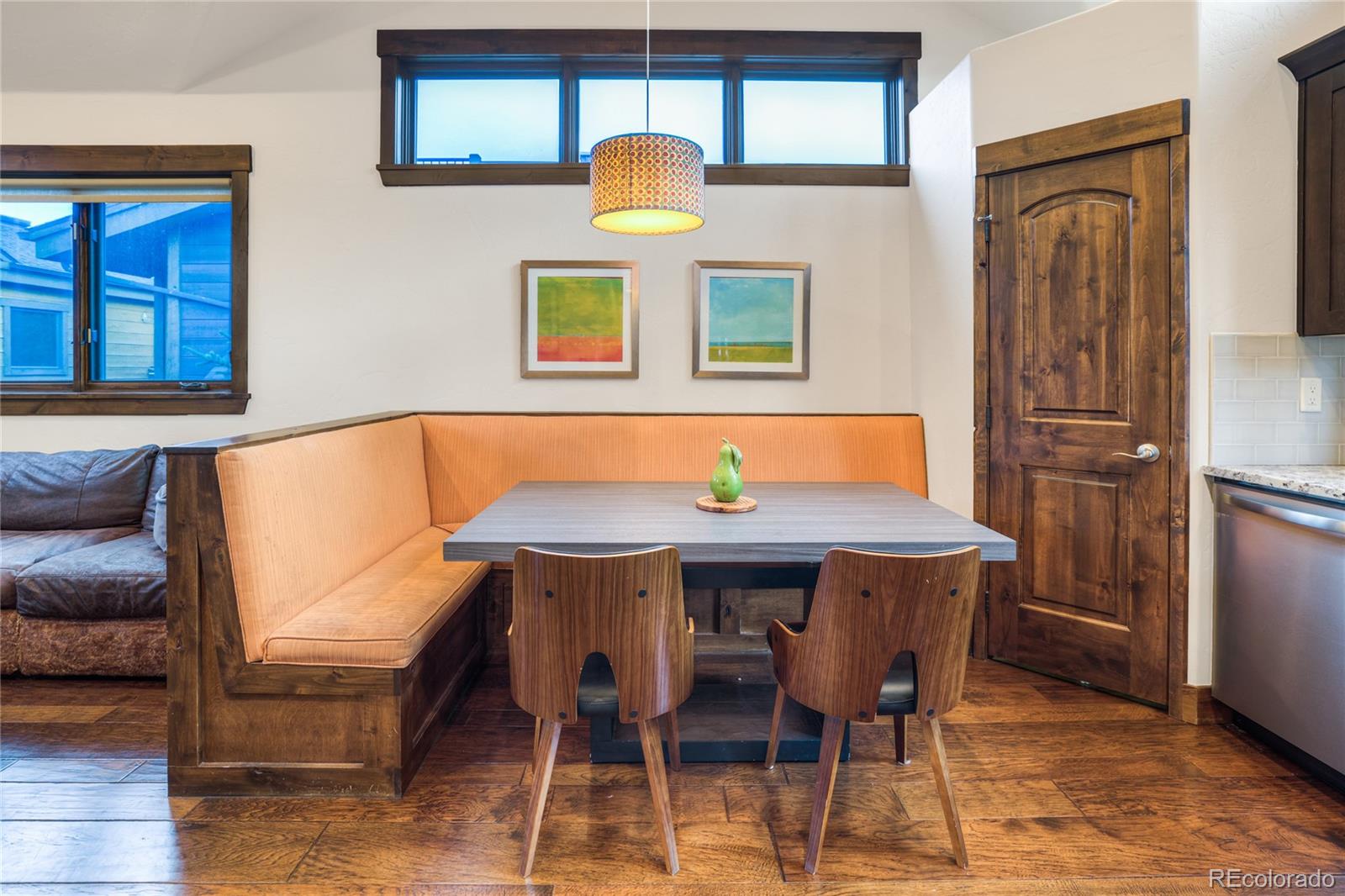 Dining nook with orange built-in bench, gray table, and two wooden chairs beneath high windows.