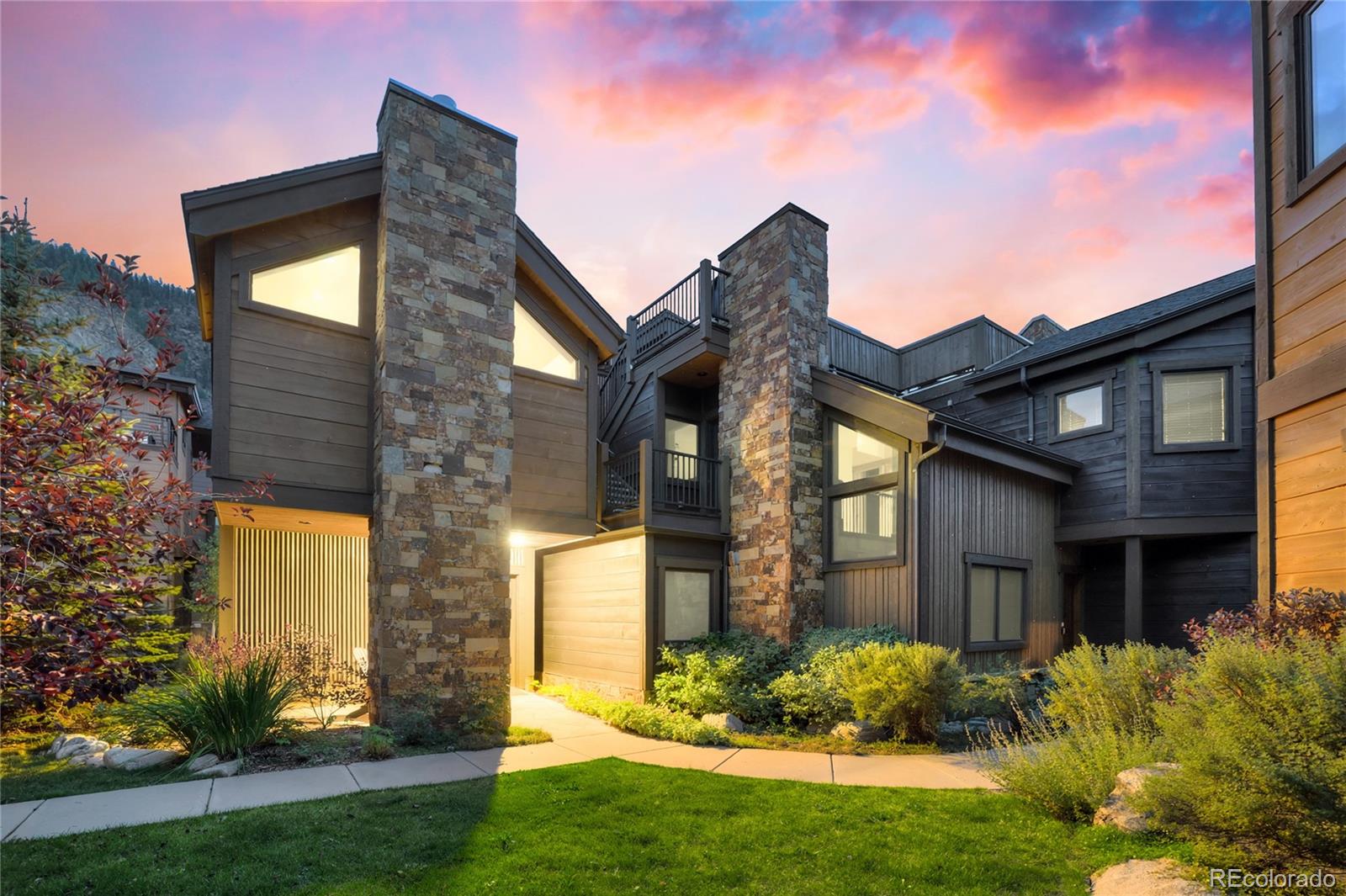 Modern mountain home with stone chimneys, dark wood siding, and landscaped grounds at sunset.