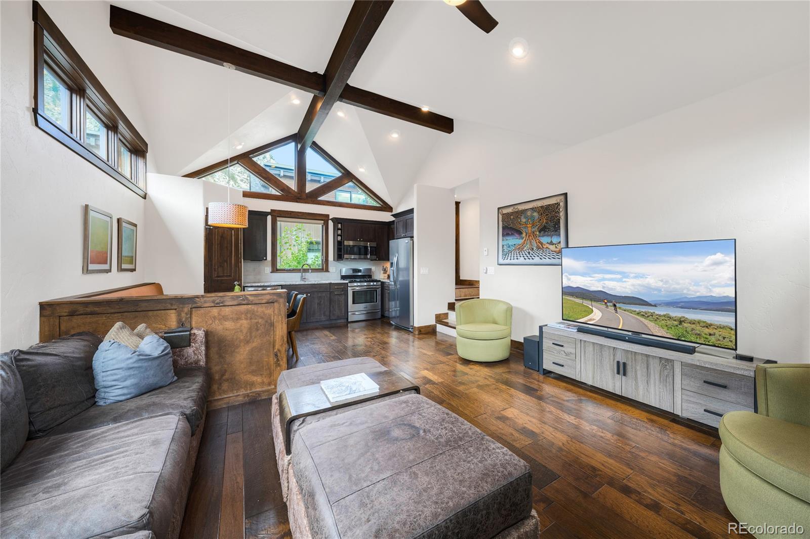Furnished living room opening to a kitchen beneath vaulted ceilings with exposed beams and triangular windows.