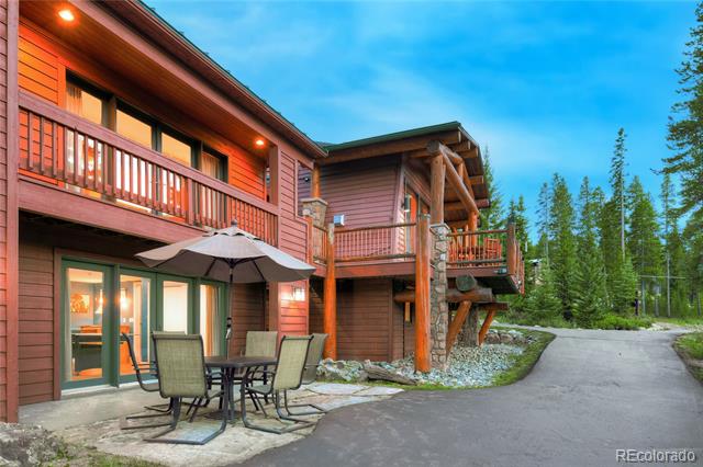 Wood-sided home with balconies, a furnished patio, and a paved driveway beside evergreen trees.