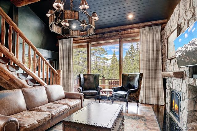 Rustic living room with leather seating, a wood staircase, stone fireplace, and large windows.