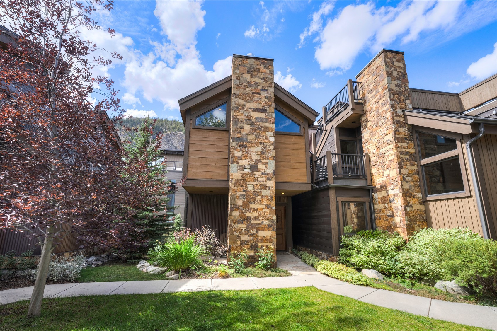 Mountain home with stone chimney columns, wood siding, and balconies amid landscaped greenery.