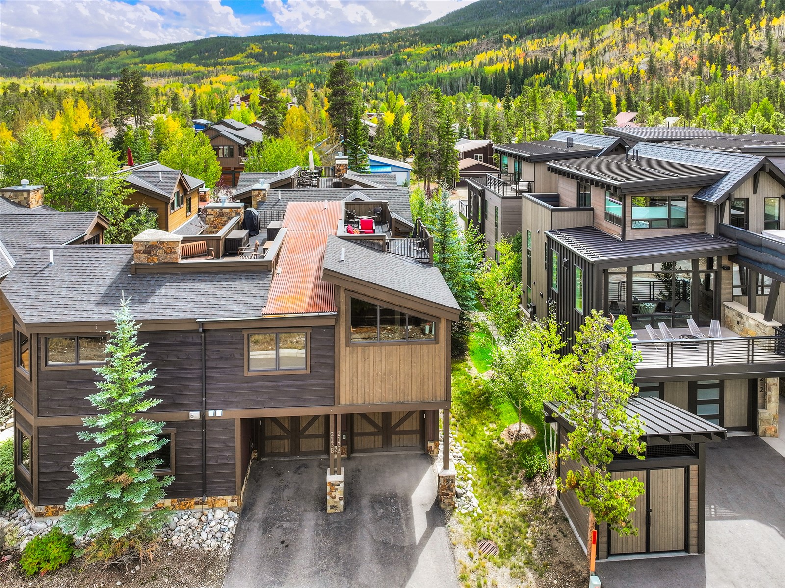 Modern dark-wood mountain home with a driveway and neighboring homes, backed by an autumn-colored forested slope.