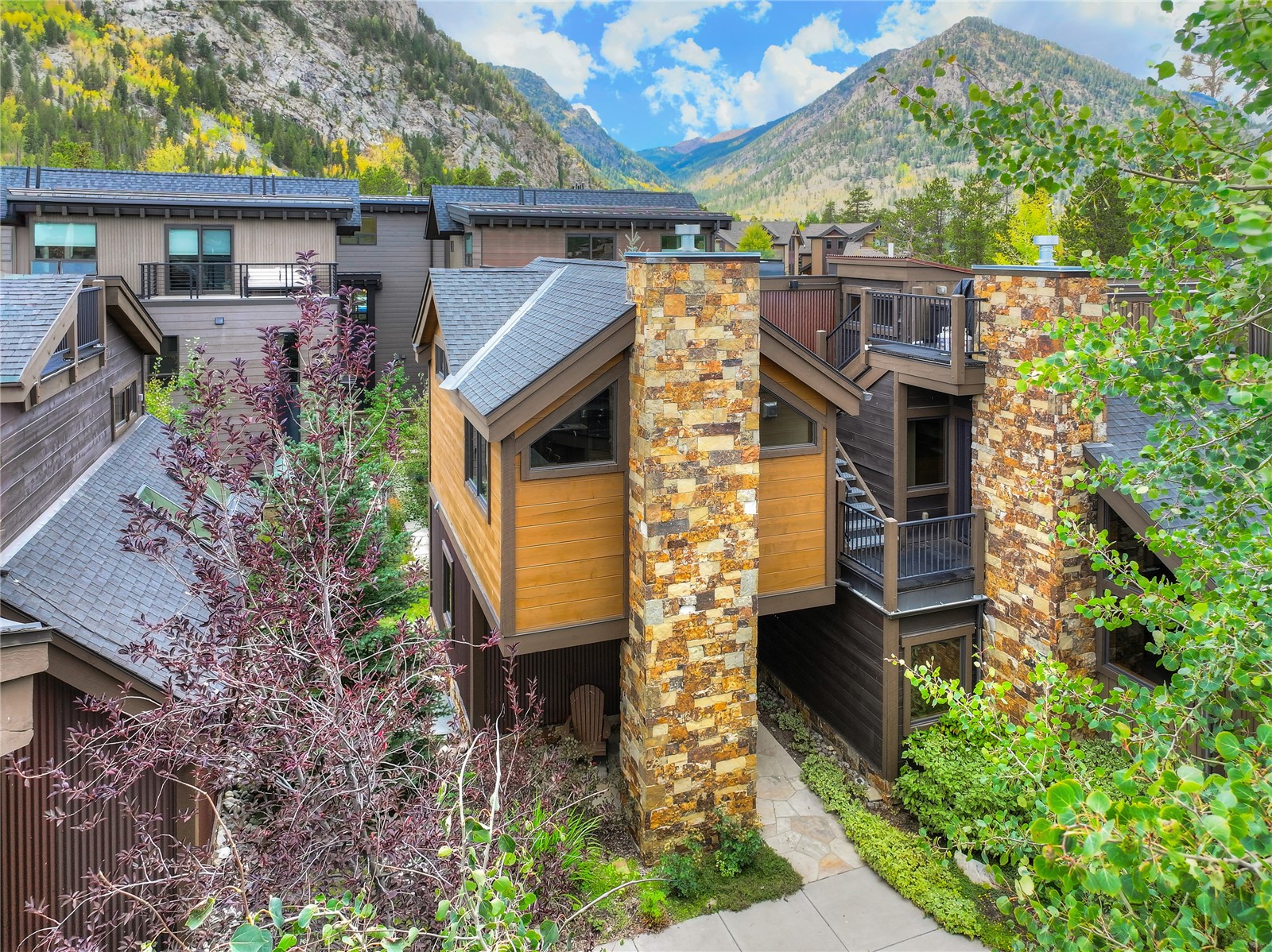 Modern mountain home with stone chimneys and balconies, framed by autumn foliage and forested peaks.