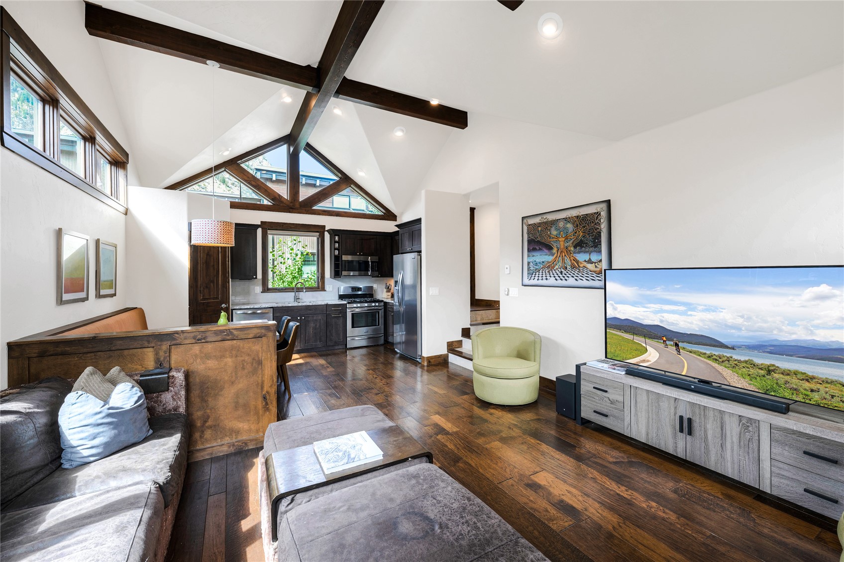 Living room with vaulted ceilings, exposed timber beams, dark wood floors, and an open kitchen.