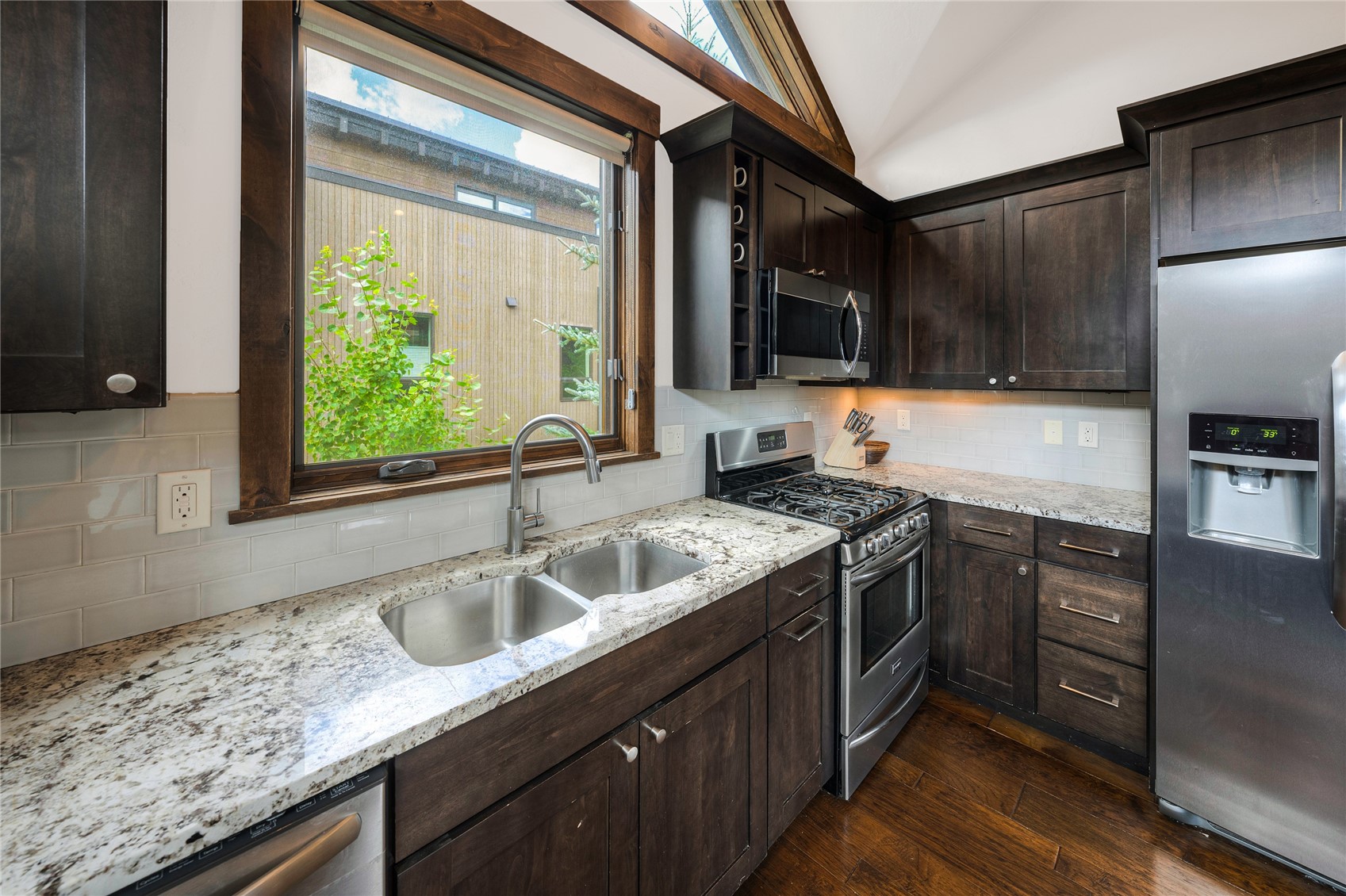 Kitchen with dark wood cabinets, granite counters, stainless steel appliances, and a double-basin sink beneath a large window.