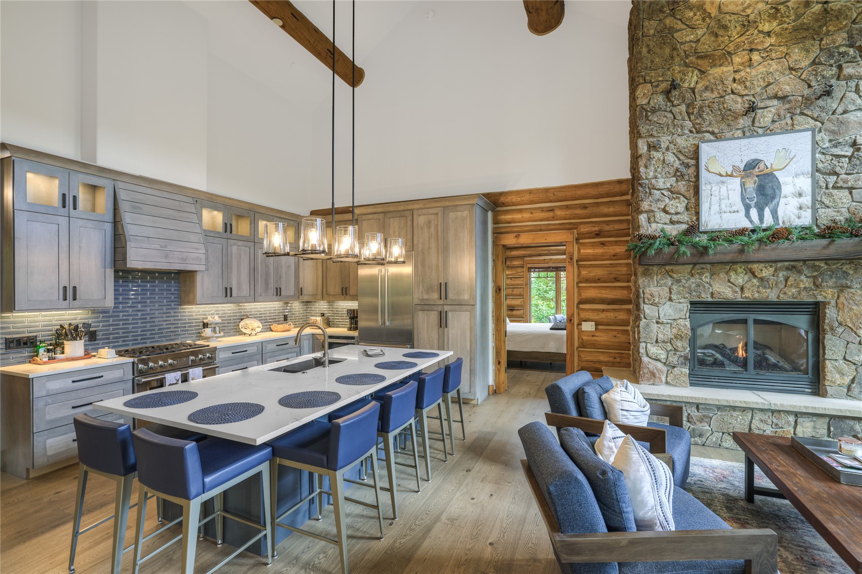 Kitchen and living area with gray cabinetry, blue island stools, and a tall stone fireplace.