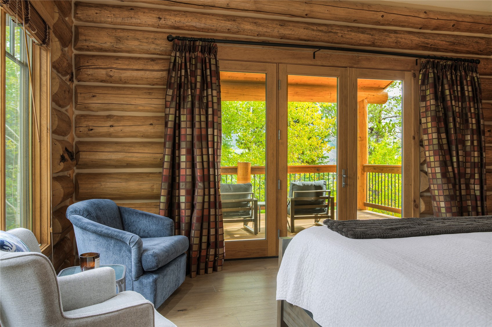 Log bedroom with exposed timber walls, a blue armchair, and glass doors to a tree-lined balcony.