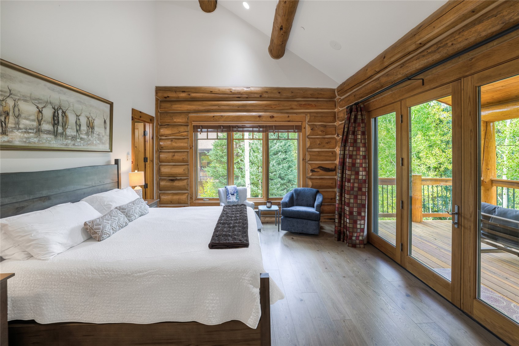 Bedroom with exposed log walls, a white bed, and glass doors opening to a leafy deck.