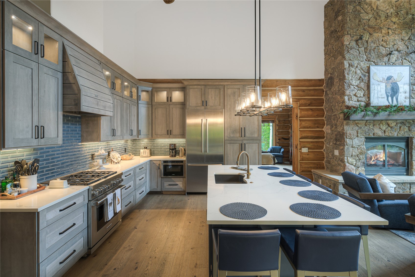 Spacious kitchen with gray cabinetry, blue tile backsplash, and a large white island beside a stone fireplace.