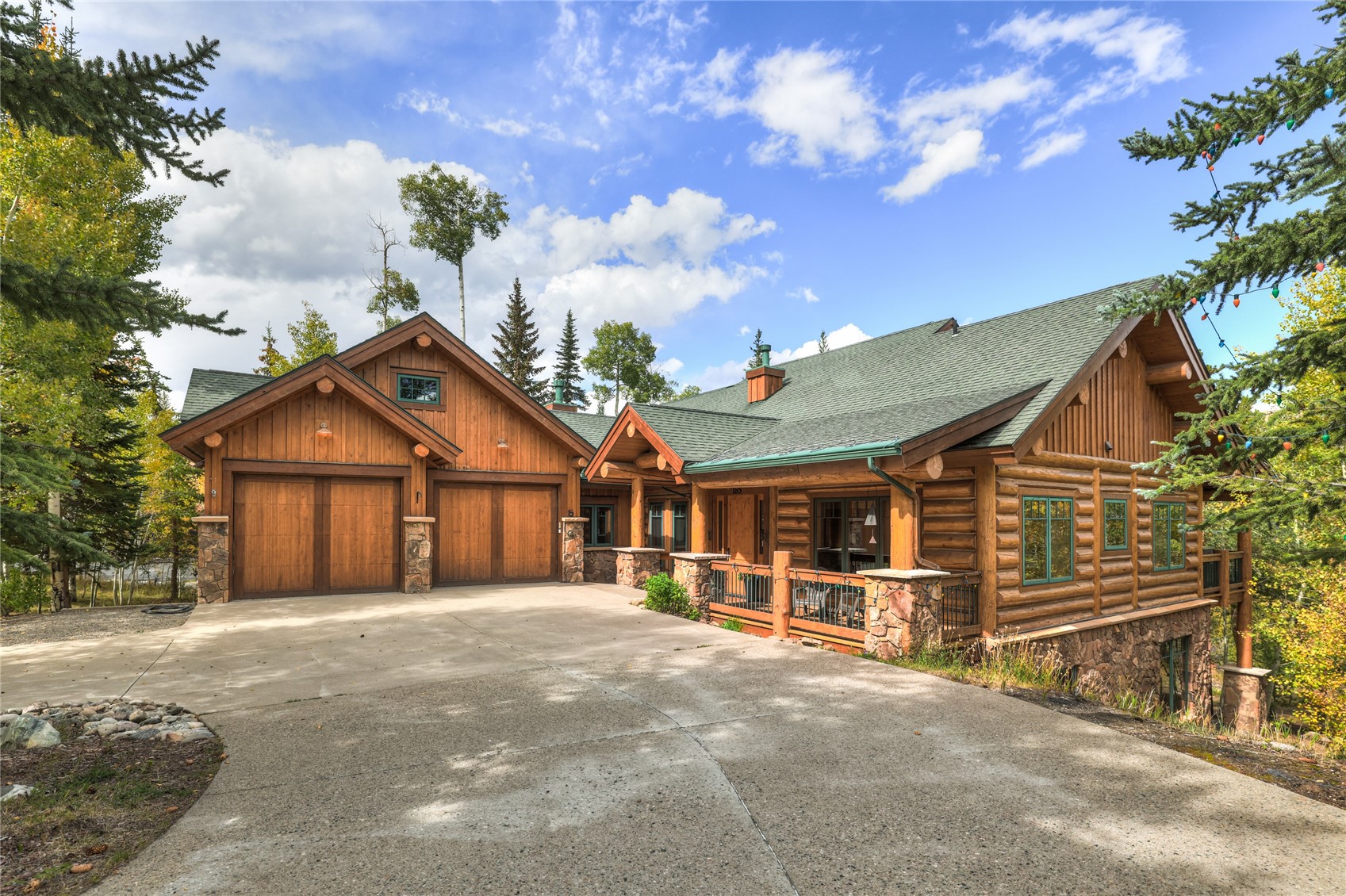 Rustic log home with a two-car garage, stone accents, and a broad concrete driveway among autumn trees.