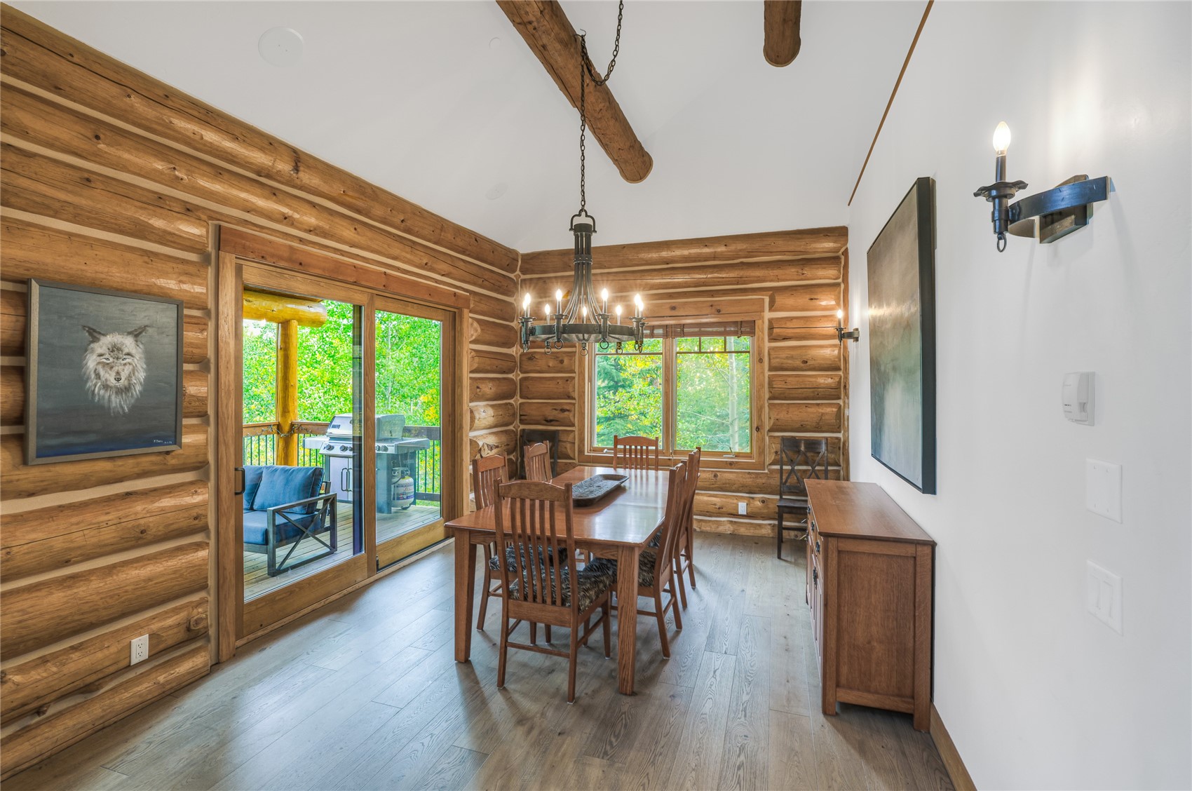 Log cabin dining room with exposed timber walls, a wooden table, and glass doors opening to a green deck.