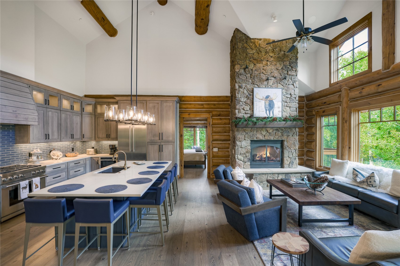 Open-plan kitchen and living room with a stone fireplace, timber beams, and a white island with blue stools.