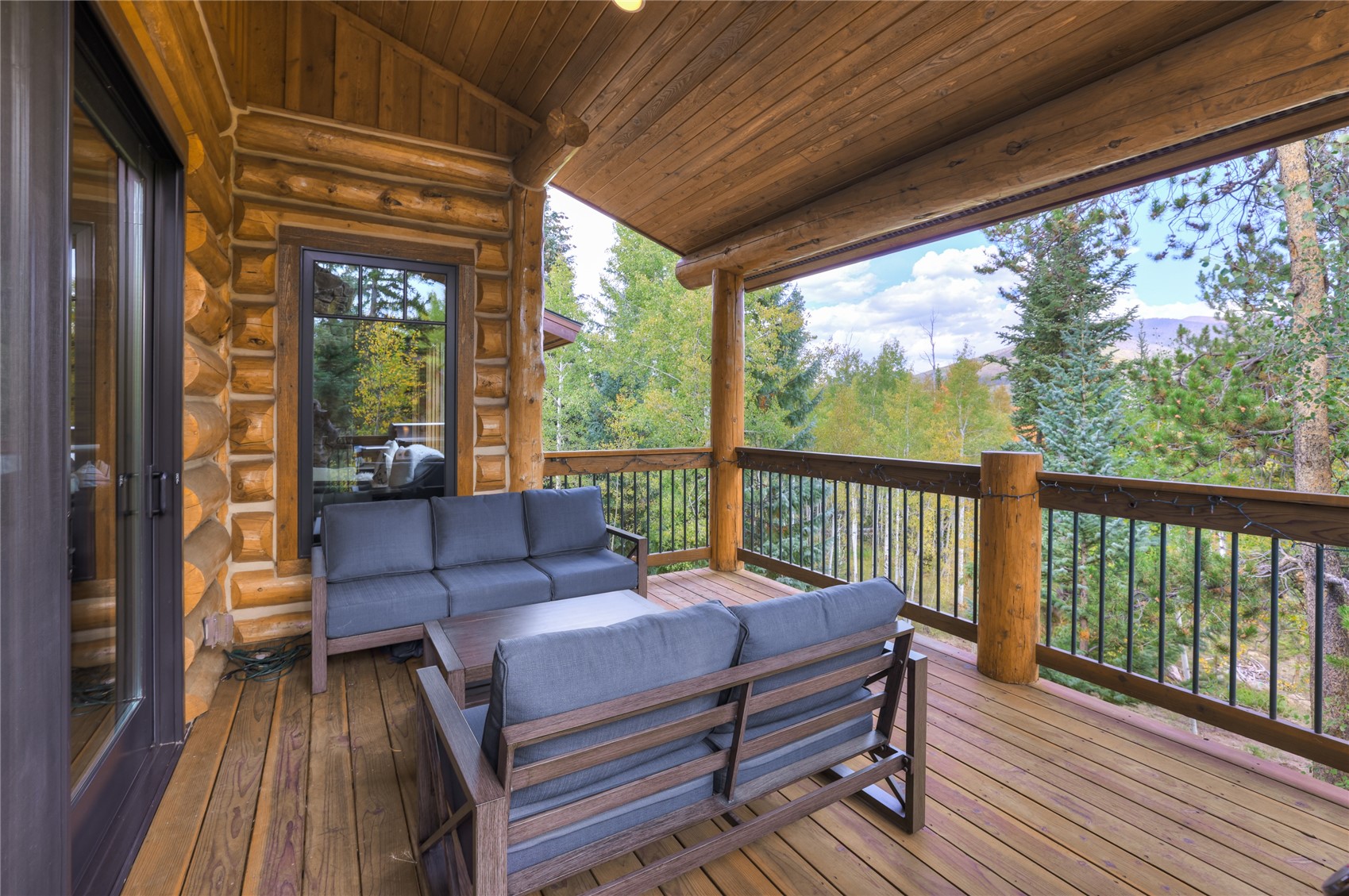 Covered log-home deck with gray outdoor seating overlooking autumn trees and a distant mountain ridge.